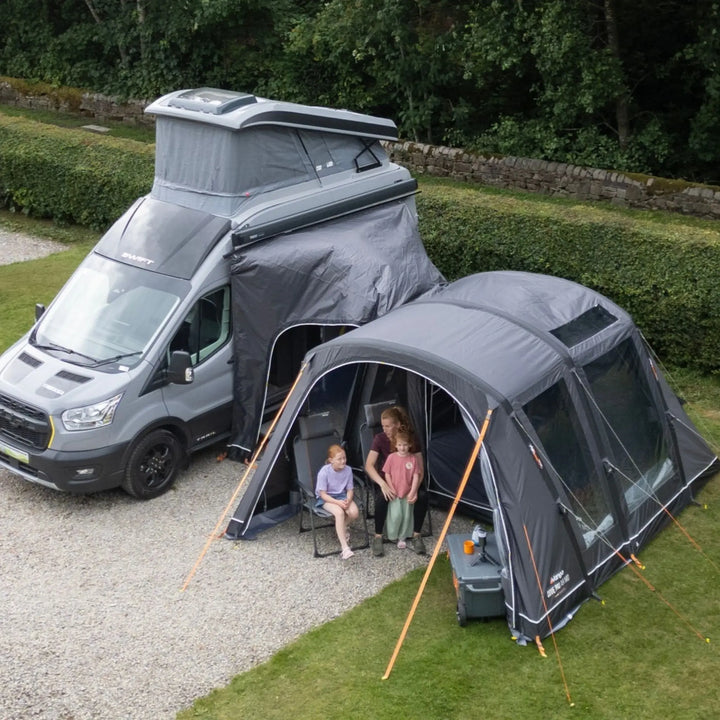 A grey pop-top camper van parked outdoors on a gravel driveway beside a Vango Cove Pro Air Mid Drive-Away Awning with two people sitting inside, with green hedges and trees in the background.