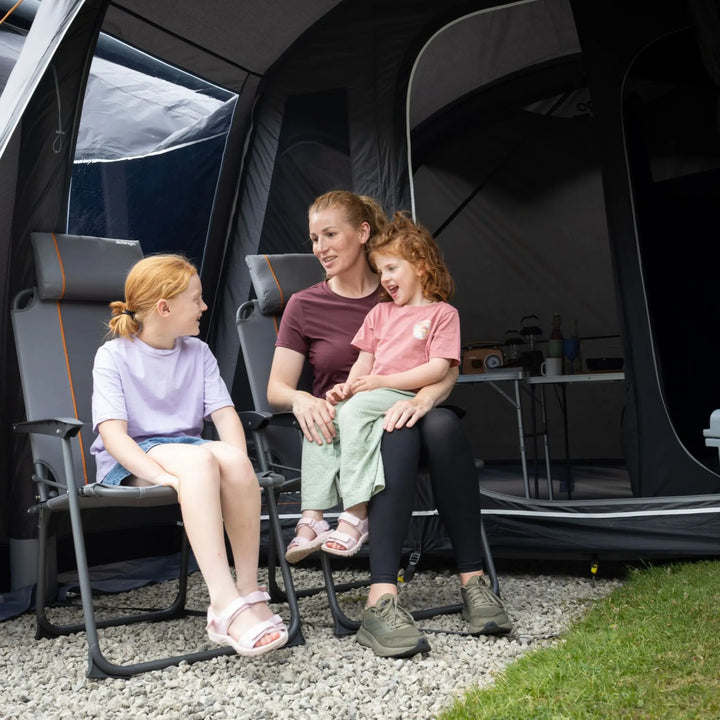 A woman and two girls sit in camping chairs inside the porch area of the Vango Cove Pro Air Mid Drive-Away Awning, with gravel and grass visible around them.