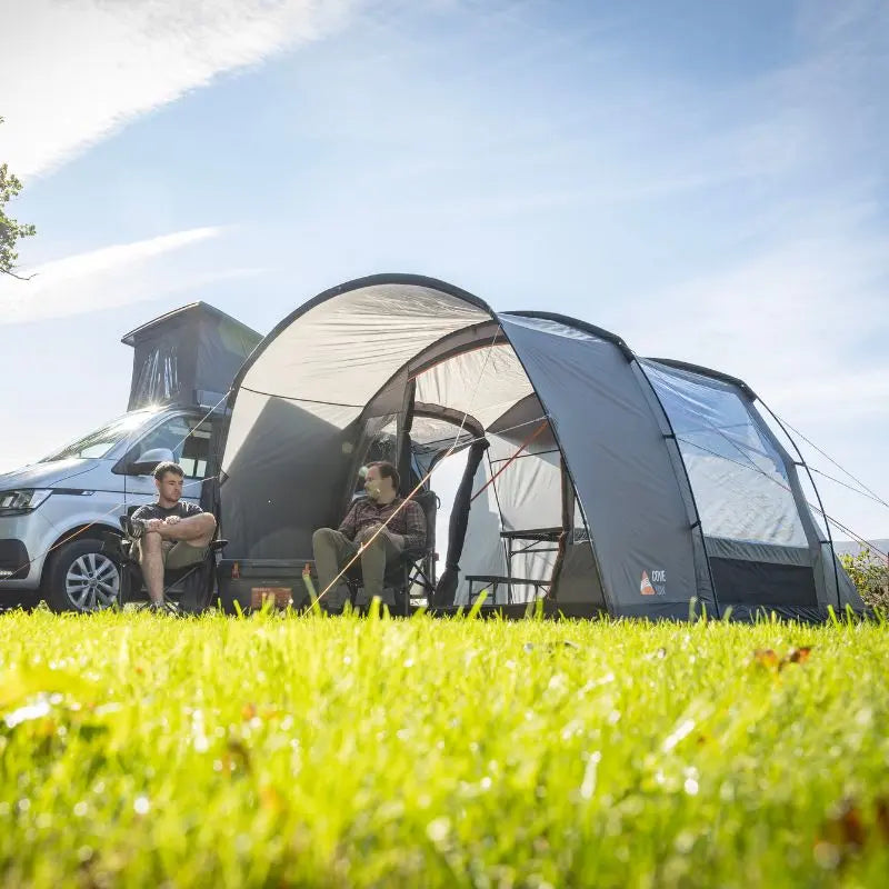 Two people sitting under a Vango Cove III Low drive away awning on a grassy area with a clear sky.