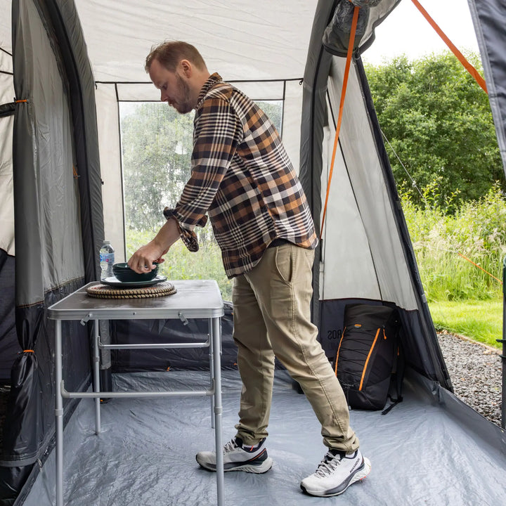 A man inside a Vango Faros III Air Mid Drive Away Awning sets a bowl on a small folding table. The awning has gray walls with orange trim, and a black backpack sits on the floor nearby.