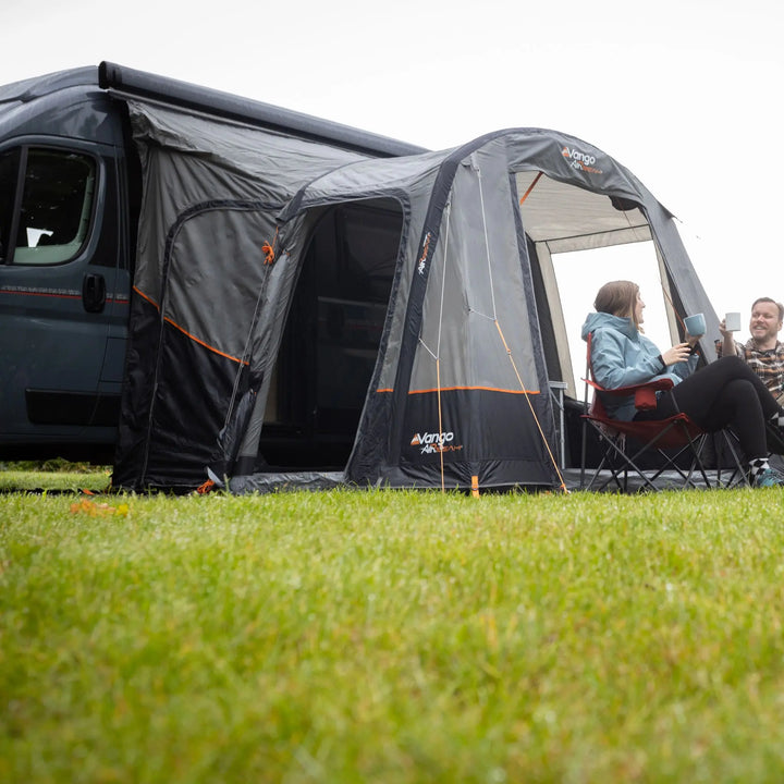 A Vango Faros III Air Mid Drive Away Awning is attached to a dark gray motorhome, set up on a green grassy field. A woman is sitting in a red folding chair next to the tent entrance, and a man is visible behind her.