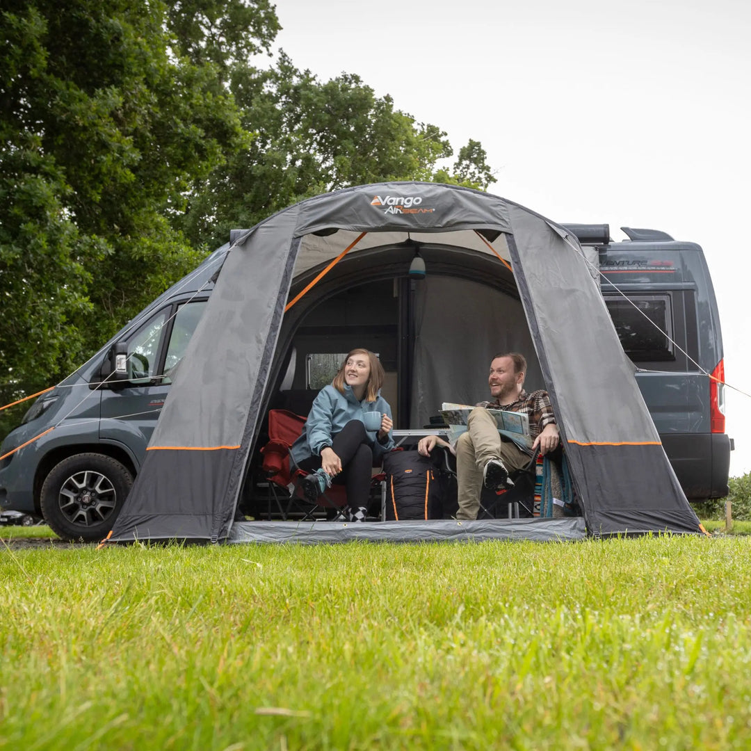 A Vango Faros III Air Mid Drive Away Awning is pitched in front of a dark camper van on a grassy lawn, with two people sitting inside the tent opening.