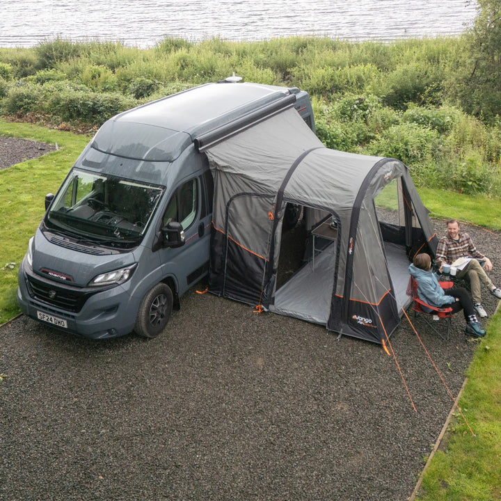 A gray motorhome parked on a gravel campsite beside a lake, with an attached Vango Faros III Air Mid Drive Away Awning. Two people are seated inside the enclosed area, and green grass and reeds are visible in the background.