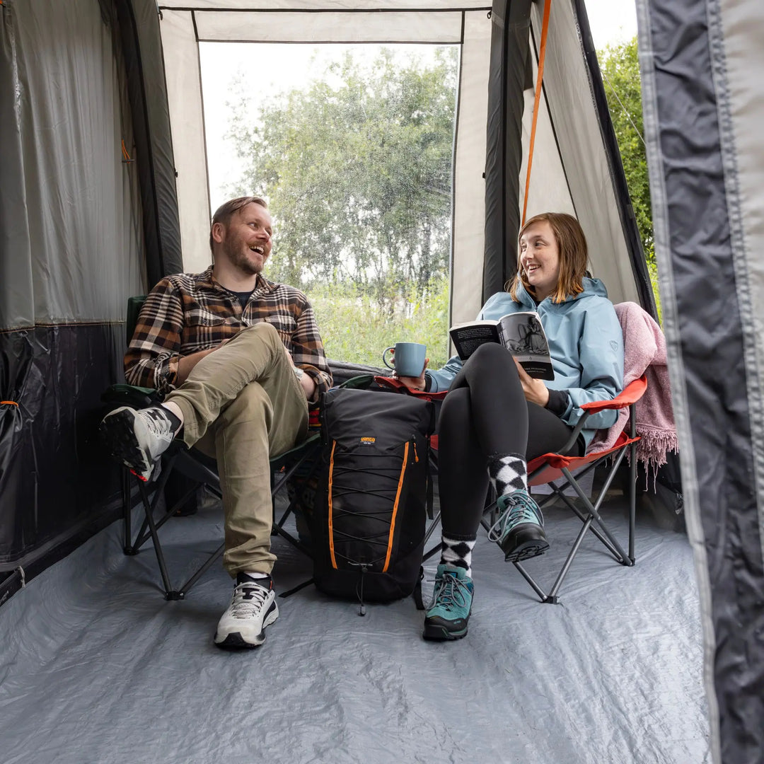 A man and a woman relax in folding camp chairs inside a Vango Faros III Air Mid Drive Away Awning, smiling and talking. The man wears a brown plaid shirt and khaki pants, while the woman wears a light blue hoodie, black leggings, and teal trainers.