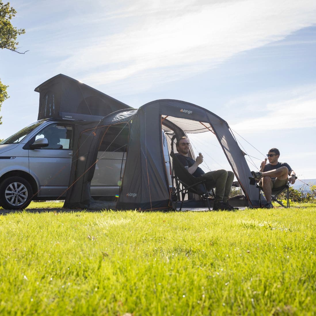 Two campers relaxing outside the Vango Faros III Low Drive Away Awning, connected to a VW campervan, in a sunny outdoor setting.