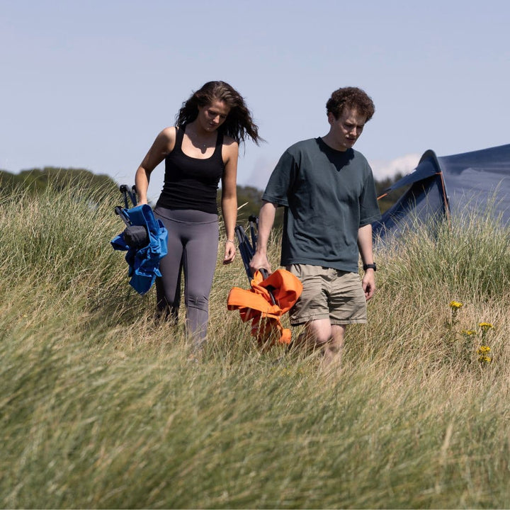 Two people walking through a grassy field carrying the Vango Fiesta Chair, demonstrating its lightweight and portable nature for outdoor adventures.