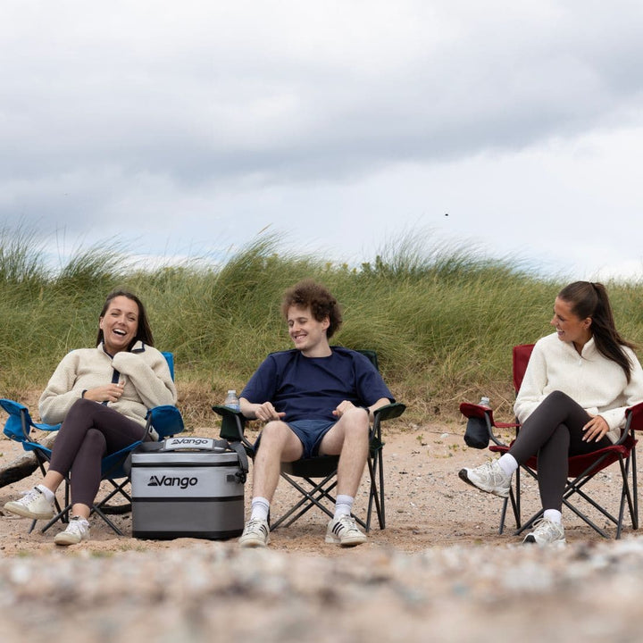 Group of friends sitting on various Vango Fiesta Chairs in cobalt and other colours on a sandy beach, enjoying the comfort of Vango camping furniture.