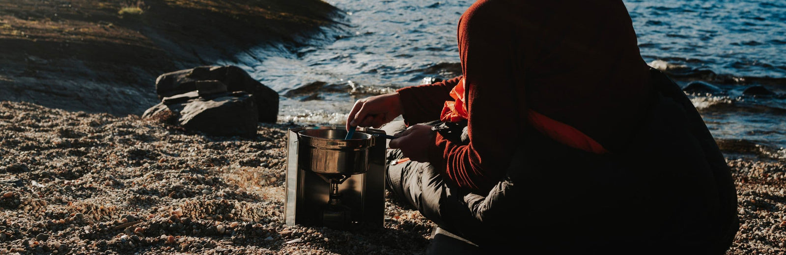 Lifestyle image of a person cooking outdoors on a rocky shore using the Vango gas stove, highlighting its suitability as a hiking stove or fishing stove for outdoor cooking in windy conditions.