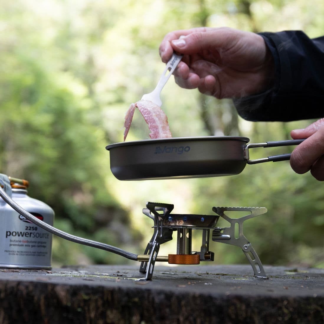 Vango Folding Gas Stove with Windshield and Piezo in use outdoors, cooking a piece of meat in a Vango pan, demonstrating its practicality as a gas stove for trekking and camping.