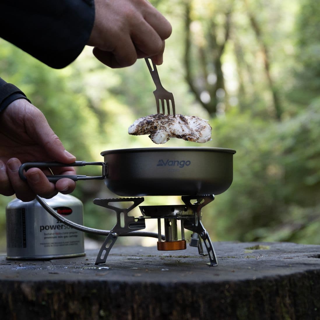 A person lifting cooked food from a Vango pan on the Vango Folding Gas Stove with Windshield and Piezo, highlighting its functionality as a portable gas stove for trekking, hiking, or fishing.