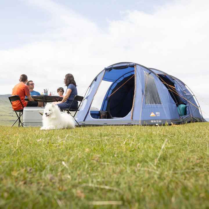 Camping scene with a Vango Iris 500 Tent, people sitting around a table, and a dog on grass.