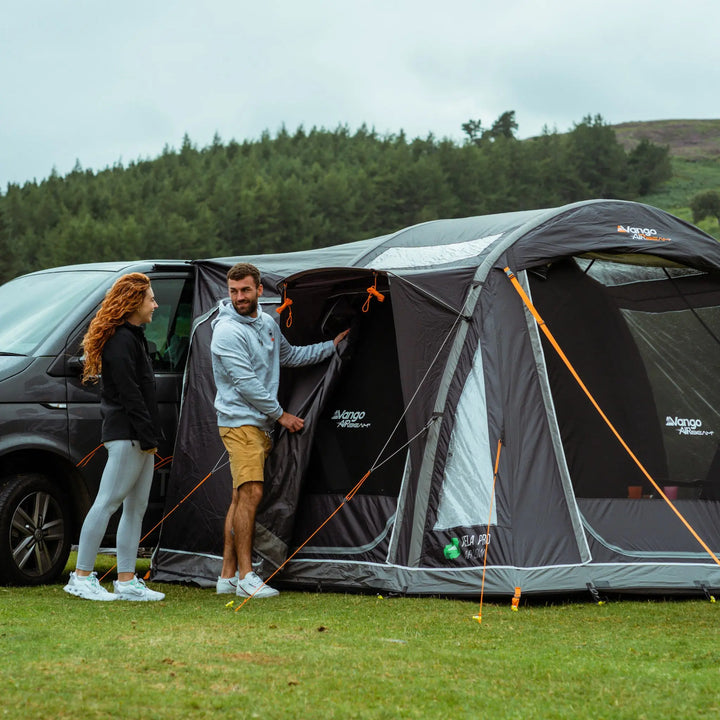 Campervan awning setup with a couple inspecting the Vango Kela Pro Air Low's side entrance, featuring orange guy ropes and zippered doors.