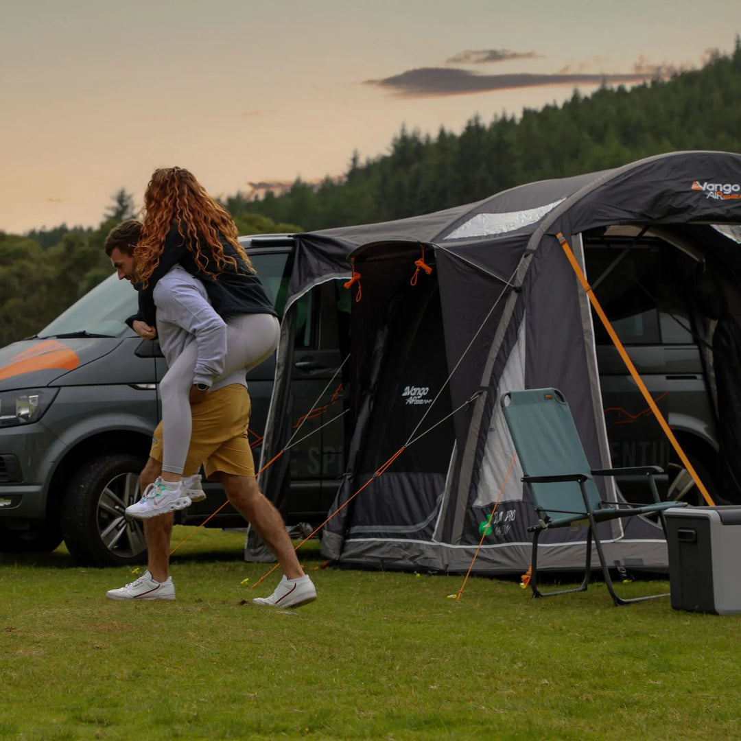 Couple enjoying a camping trip with the Vango Kela Pro Air Low campervan awning, with outdoor Vango chairs and a portable cooler set up nearby.