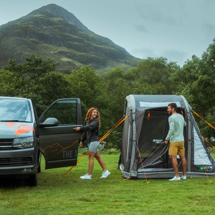 Vango Kela Pro Air Low campervan awning pitched in a scenic mountain location, with a couple preparing to enter the awning while parked next to a campervan.