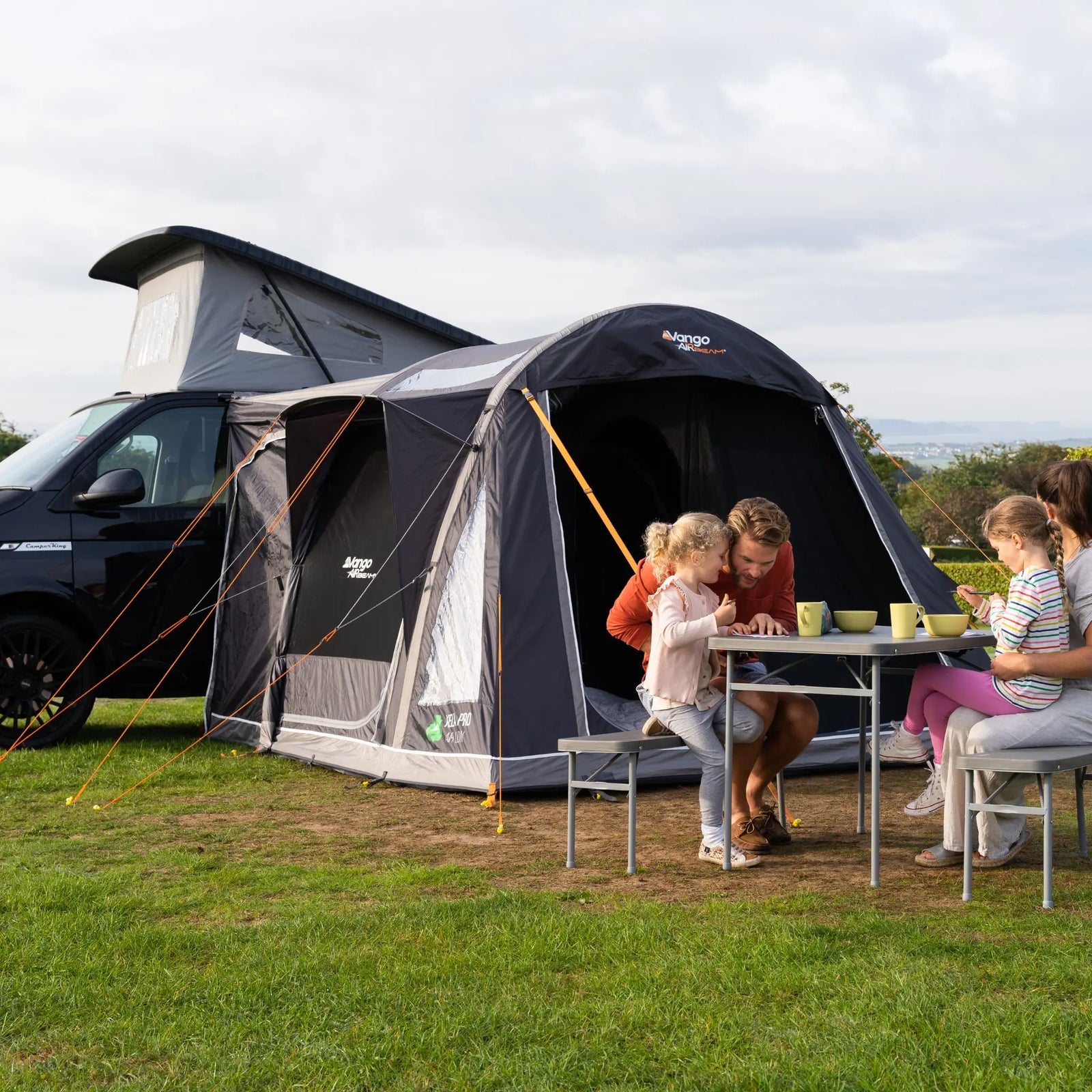 Family enjoying an outdoor meal beside the Vango Kela Pro Air Low campervan awning, with a pop-top campervan parked nearby.