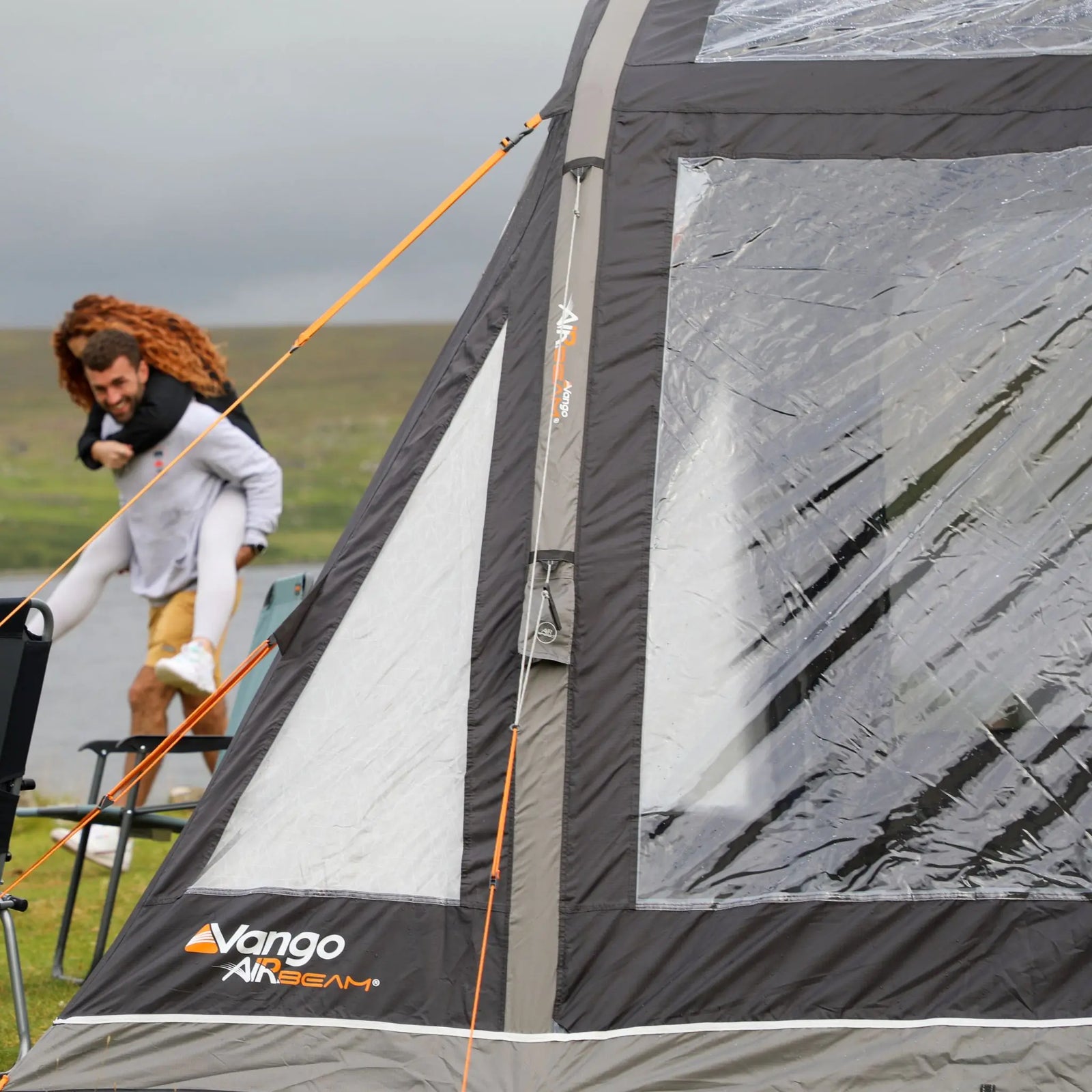 Close-up of the Vango AirBeam campervan awning, showcasing large clear windows and tensioned guy ropes, with a couple playing in the background.