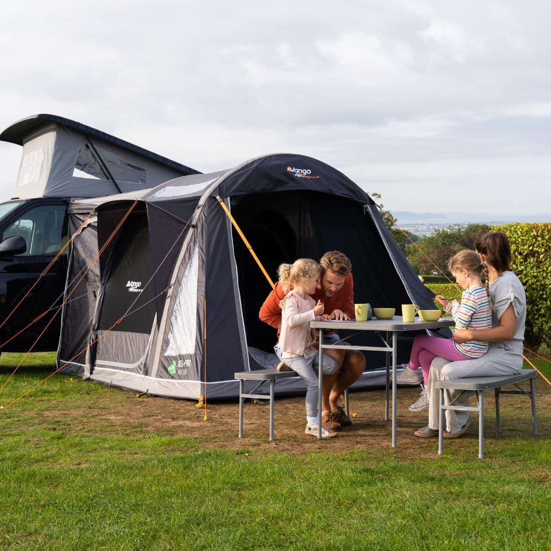 Family sitting at a table outside the Vango Kela Pro Air Low Drive Away Awning, demonstrating the versatile use of this low-height inflatable campervan awning for camping trips.