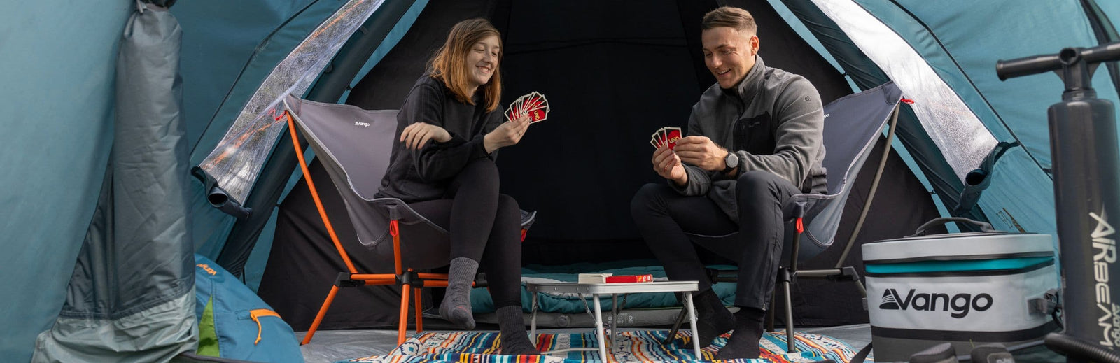 Two campers sitting on Vango Micro Lux Chairs (Excalibur) inside a tent, enjoying a game of cards with camping accessories around them.