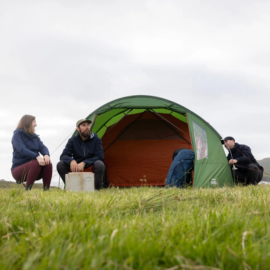 A Vango Omega 600xl Tent with an orange interior pitched in a grassy field under an overcast sky, with three people sitting and crouching around it.