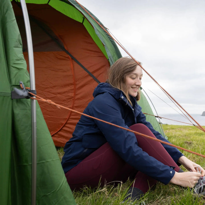 A woman sits on the grass in front of a Vango Omega 600xl Tent, tying her shoe outdoors under an overcast sky.