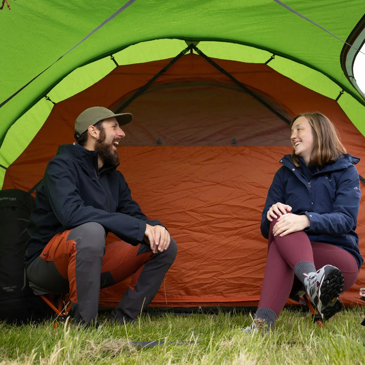 A man and a woman sitting inside a Vango Omega 600xl Tent on a grassy field, smiling and talking.
