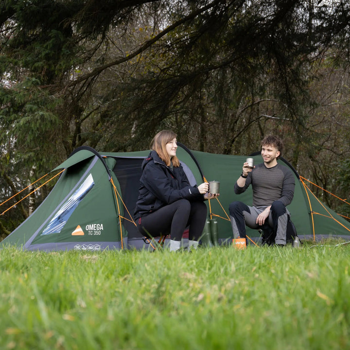 A couple sits in front of a green Omega TC 350 camping tent in a grassy forest clearing, holding mugs and cups.