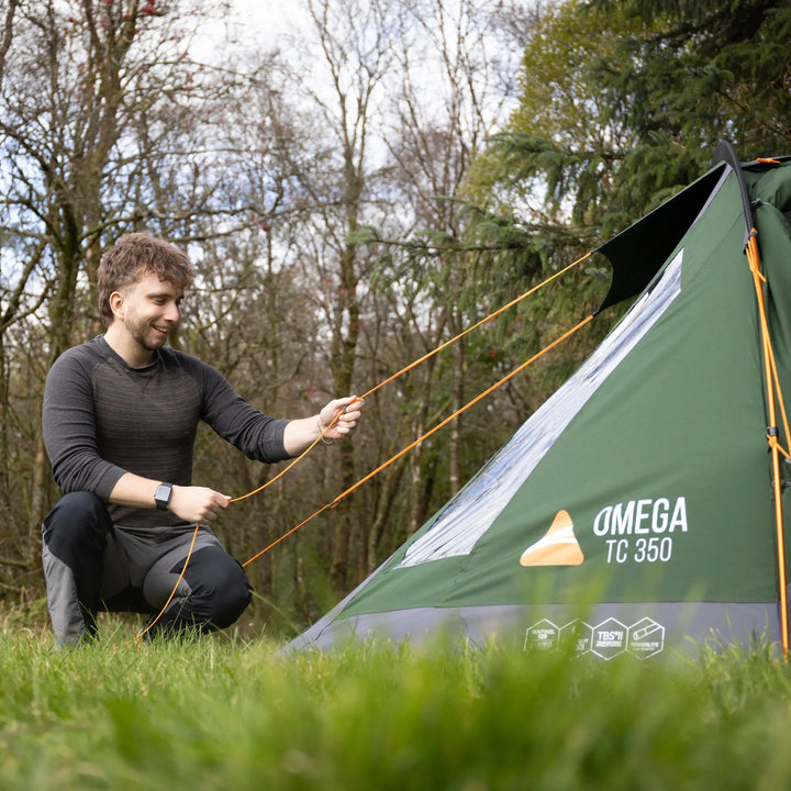A man kneeling in a grassy outdoor area sets up an OMEGA TC 350 camping tent, adjusting orange guy lines against a backdrop of trees.