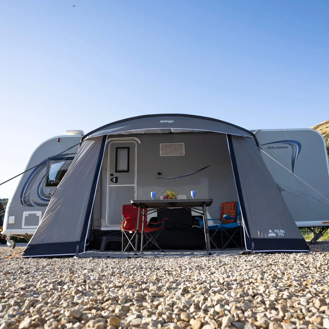 A Vango Palma 330 Poled Caravan Awning is set up on a gravel campsite in front of a white caravan. The awning is open to reveal a small dining table, chairs, and a bed inside, with the blue sky behind it.