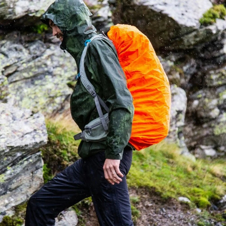 Hiker wearing a waterproof jacket and a 60+ litre rucksack protected by the bright orange Vango Rain Cover - Large, walking through wet, rocky terrain in the rain.