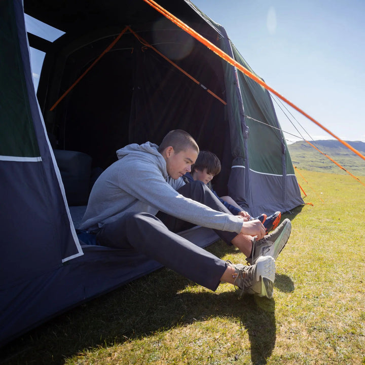 A man and a child sit inside an open Vango Sherwood Air TC 400 Tent in a grassy field, with the man helping the child with a shoe. The tent is dark green and navy blue with orange guy lines, under a clear blue sky.