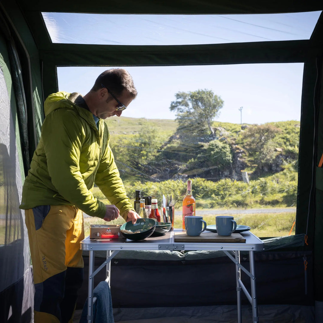 A man in a green hooded jacket and yellow pants sets up a small folding table inside a Vango Sherwood Air TC 400 Tent, with mugs, bottles, and outdoor scenery visible through the open back door.