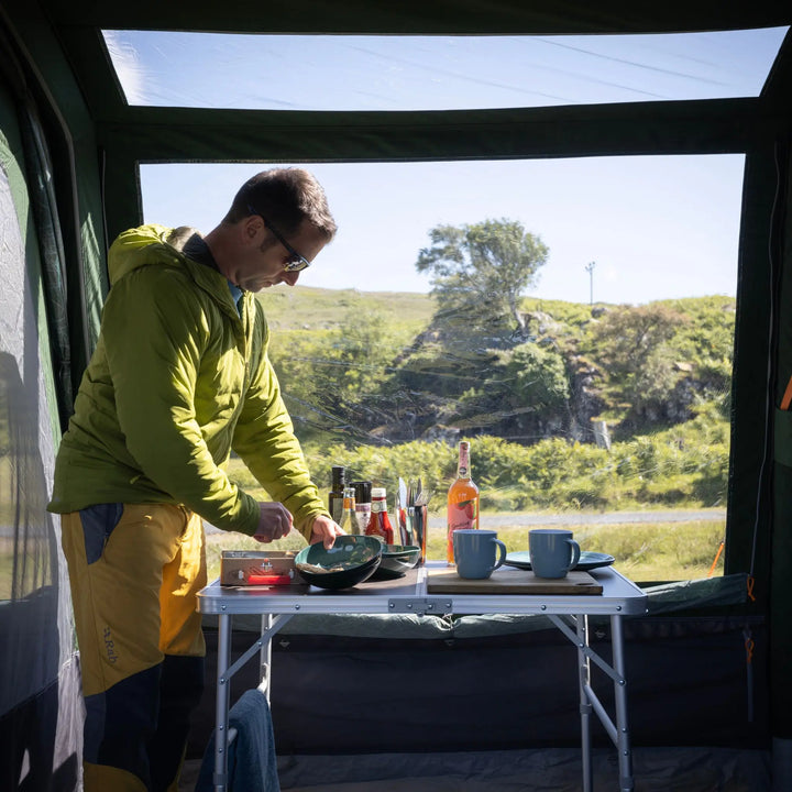 A man in a green hooded jacket and yellow pants sets up a small folding table inside a Vango Sherwood Air TC 400 Tent, with mugs, bottles, and outdoor scenery visible through the open back door.