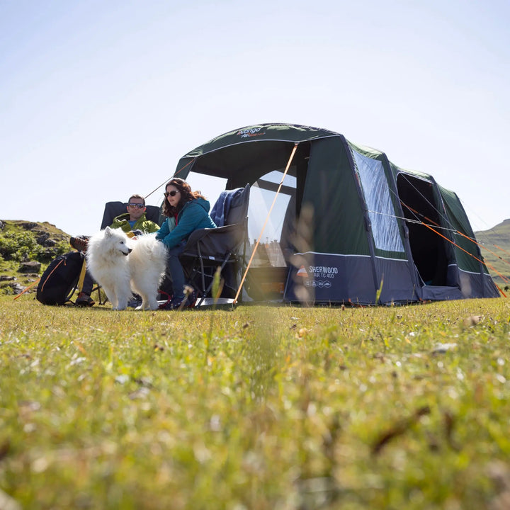 A Vango Sherwood Air TC 400 Tent pitched on grassy ground with a woman and two dogs sitting beside it under a clear sky.