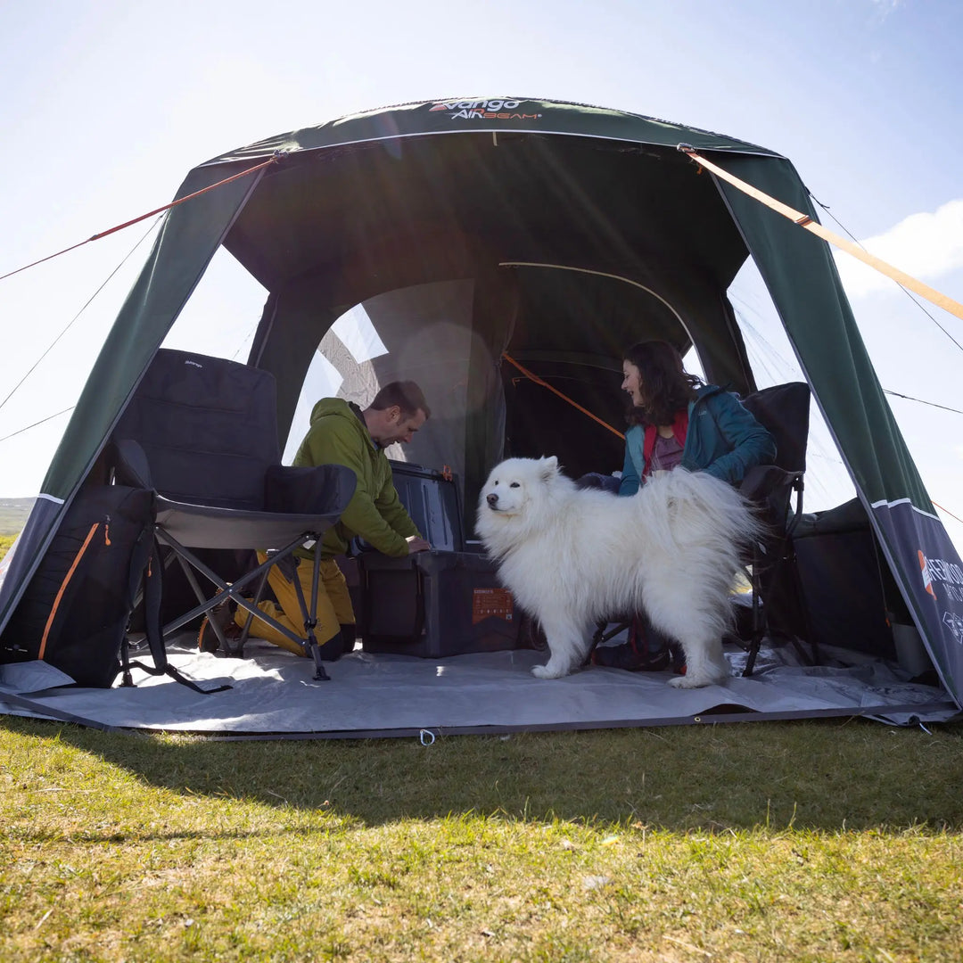 A Vango Sherwood Air TC 400 Tent set up outdoors on grass, with two people sitting inside and a fluffy white dog standing in front of the entrance. The tent has visible Vango branding on the front and side panels.