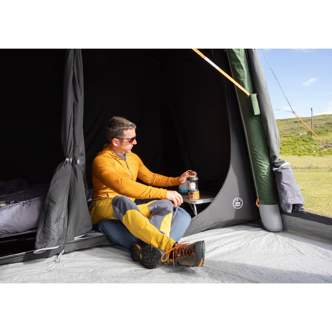 A man wearing a yellow and blue camping jacket sits inside a Vango Sherwood Air TC 400 Tent, holding a lantern. The tent is set in an outdoor grassy field under a blue sky.
