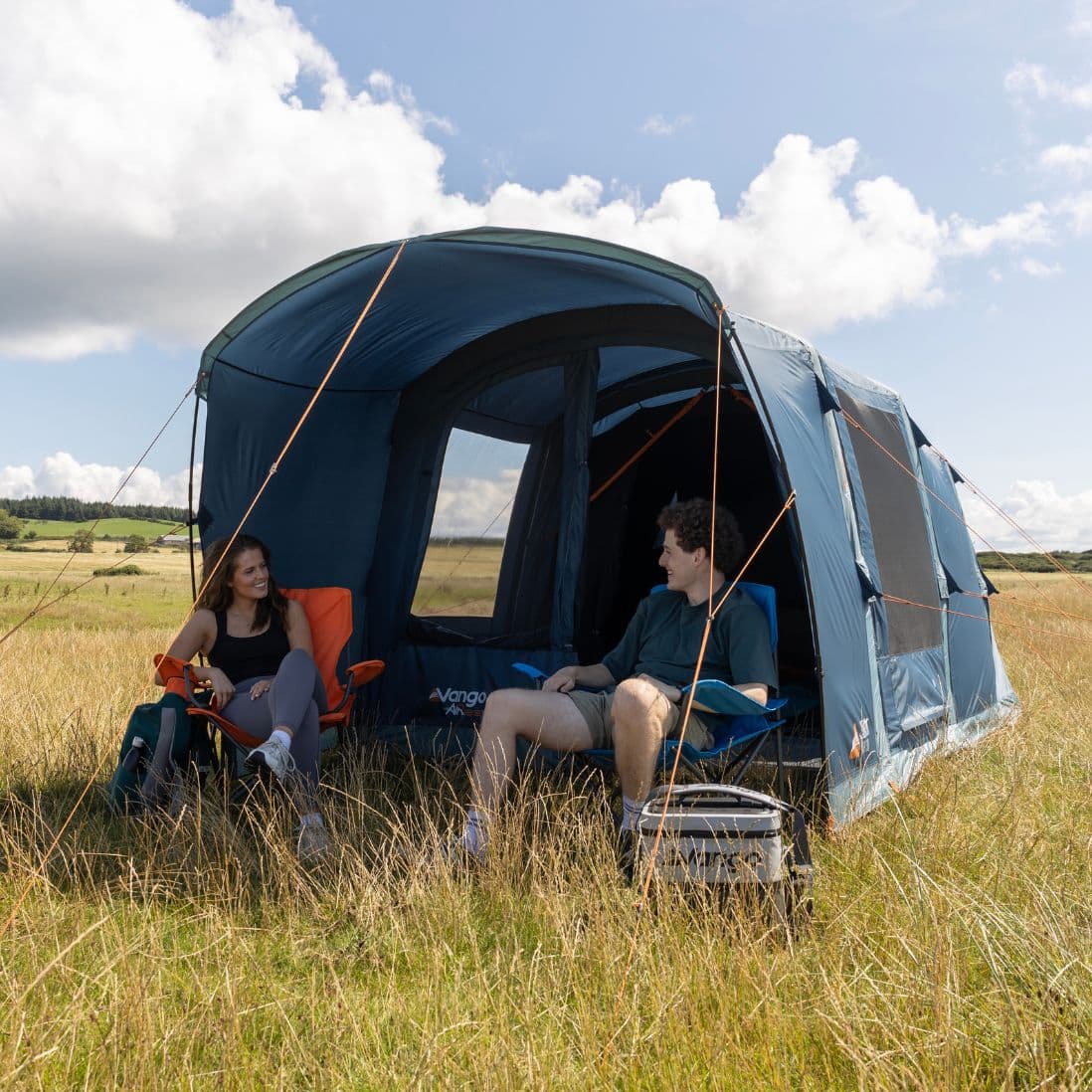 Two campers seated inside the Vango Sierra Air 300 couples tent, enjoying the open front and views of the surrounding grassy landscape.