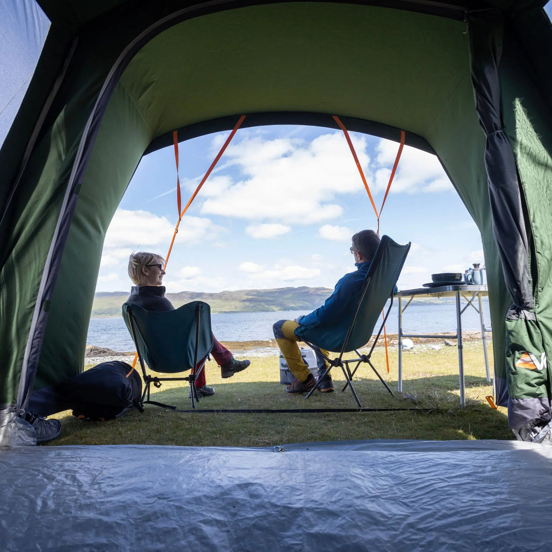 Two people sitting outside a Vango Sierra TC 300 Poled Tent overlooking a scenic view of water and hills.