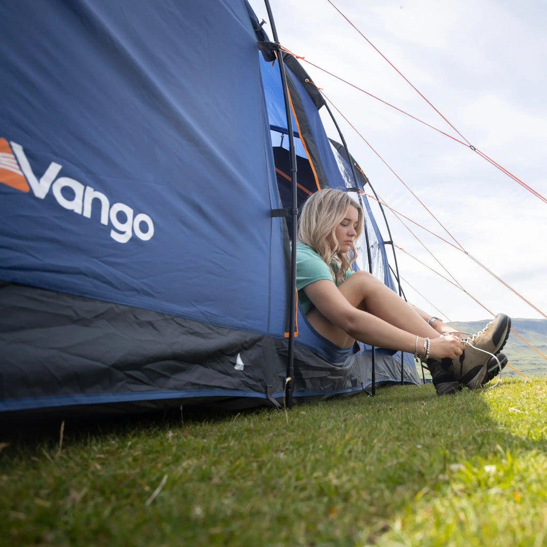 Woman sitting outside a Vango Sorrento 600XL Tent in a grassy area with mountains in the background
