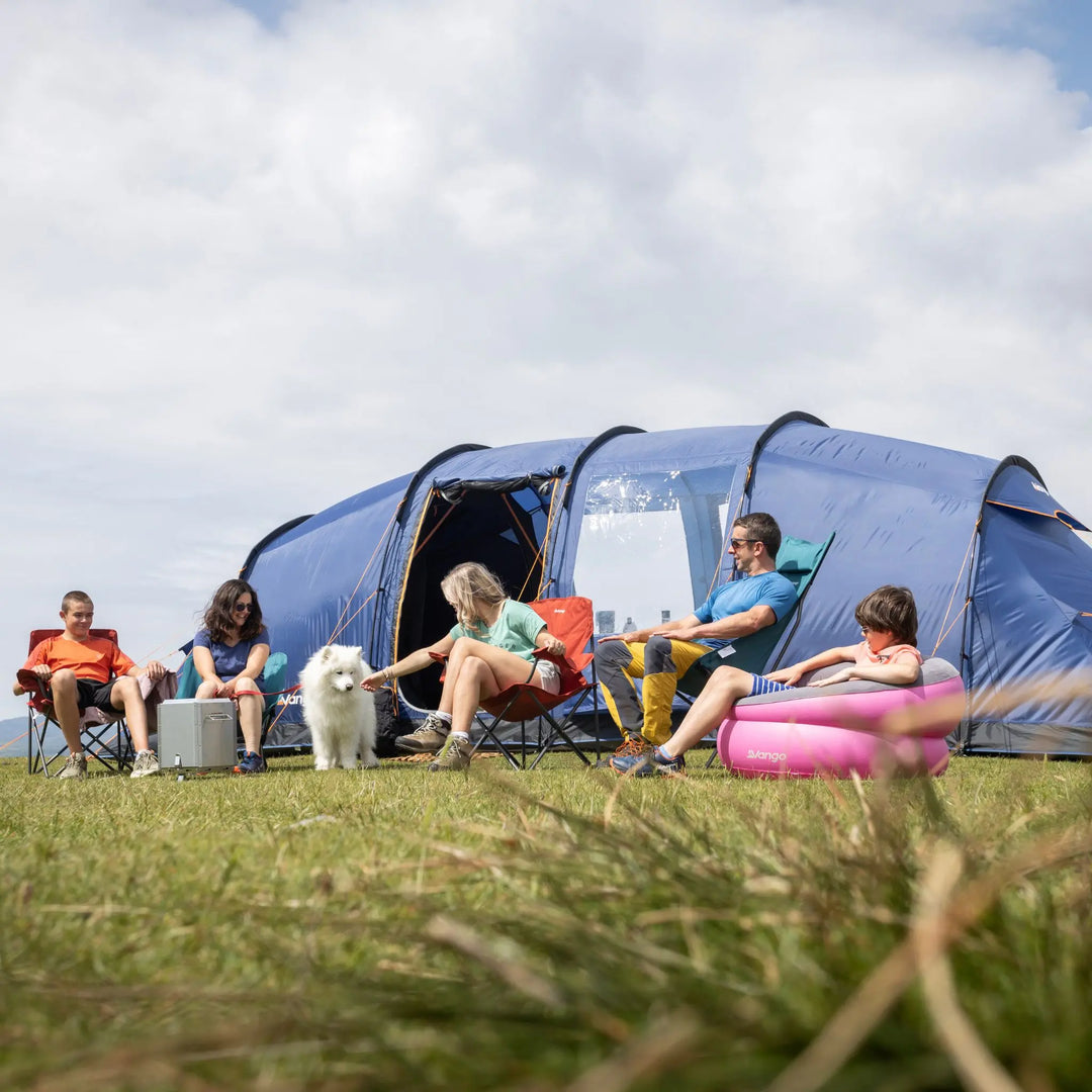 Family with a dog sitting outside a Vango Sorrento 600XL Tent in a grassy area under a cloudy sky.