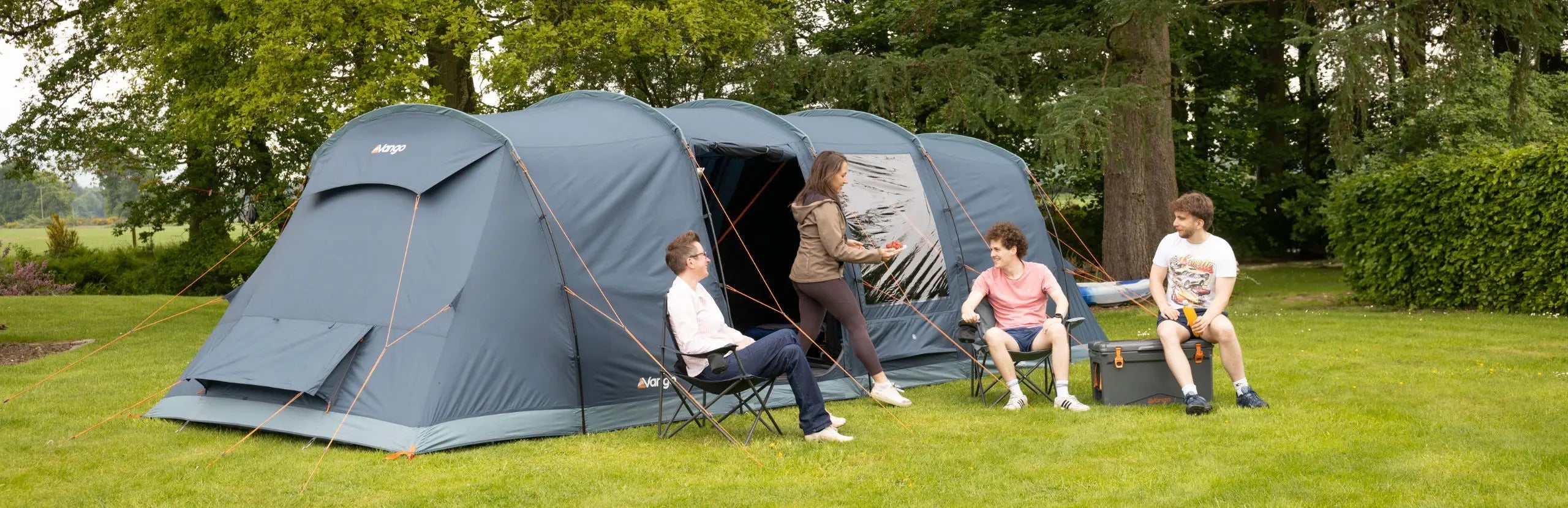 Vango Tacoma 800xl family tent pitched on a green lawn with trees in the background. Several people are sitting and standing around the tent, using folding chairs and standing near the entrance.