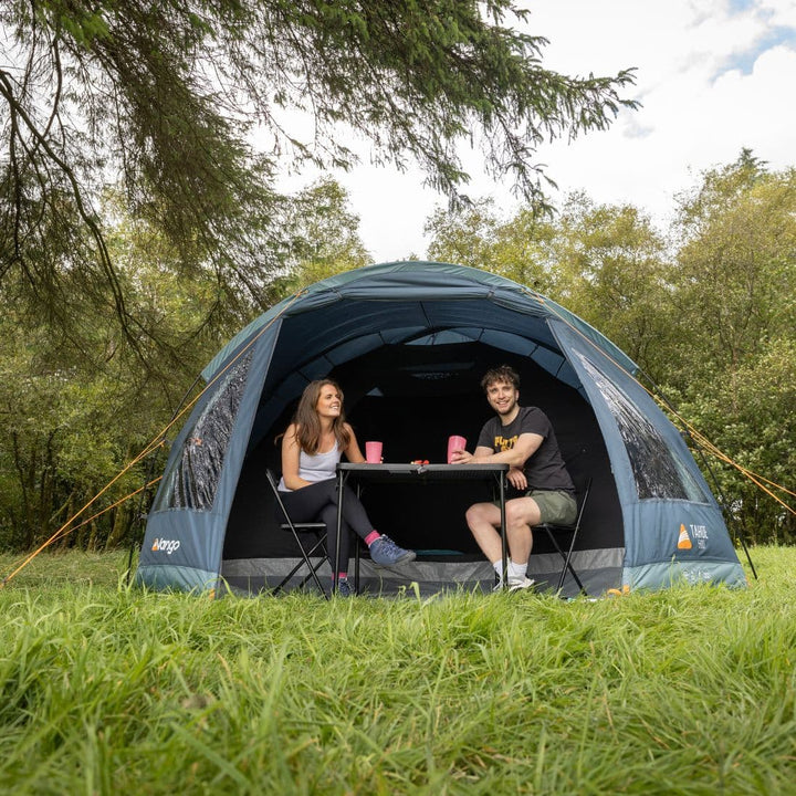 A Vango Tahoe 500 5-man tent set up in a scenic outdoor setting, with a couple enjoying a meal at the entrance. Perfect for festivals and weekend camping