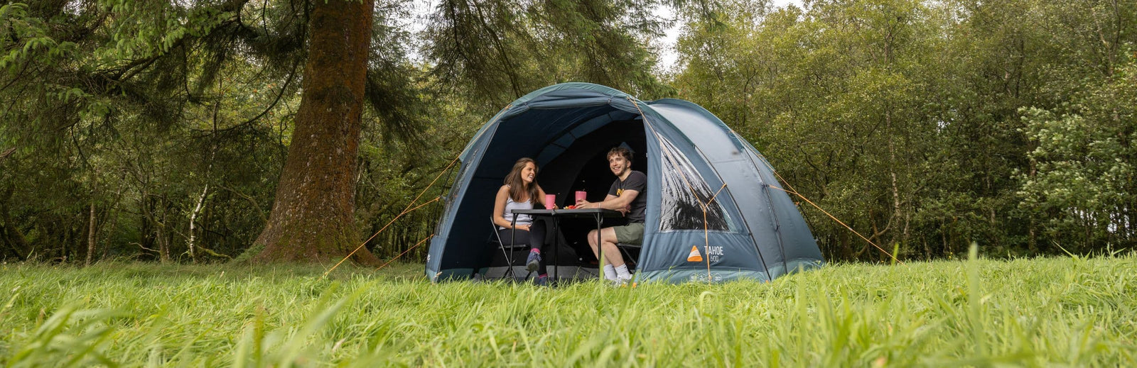 The Vango Tahoe 500 tent in a lush green setting, with a couple seated inside, enjoying its comfort and spacious design. Ideal for weekend trips or festivals