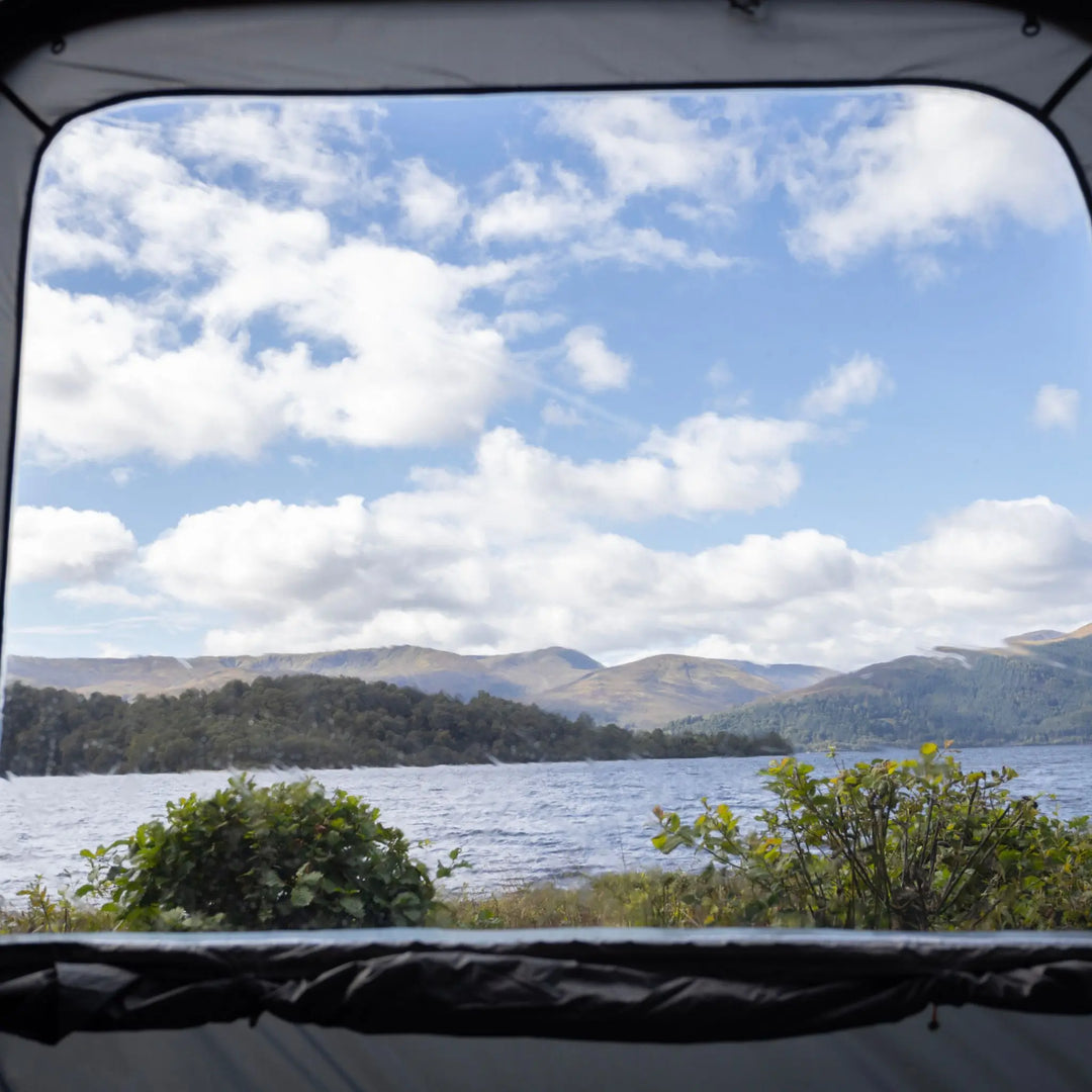 Scenic lake and mountain view framed through the rear window of the Vango Tailgate Hub II Low awning, highlighting its diamond clear windows.