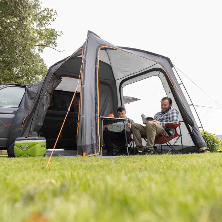 Front view of the Vango Tailgate Hub II Low awning, with two people relaxing on camping chairs, highlighting the liveable space inside.