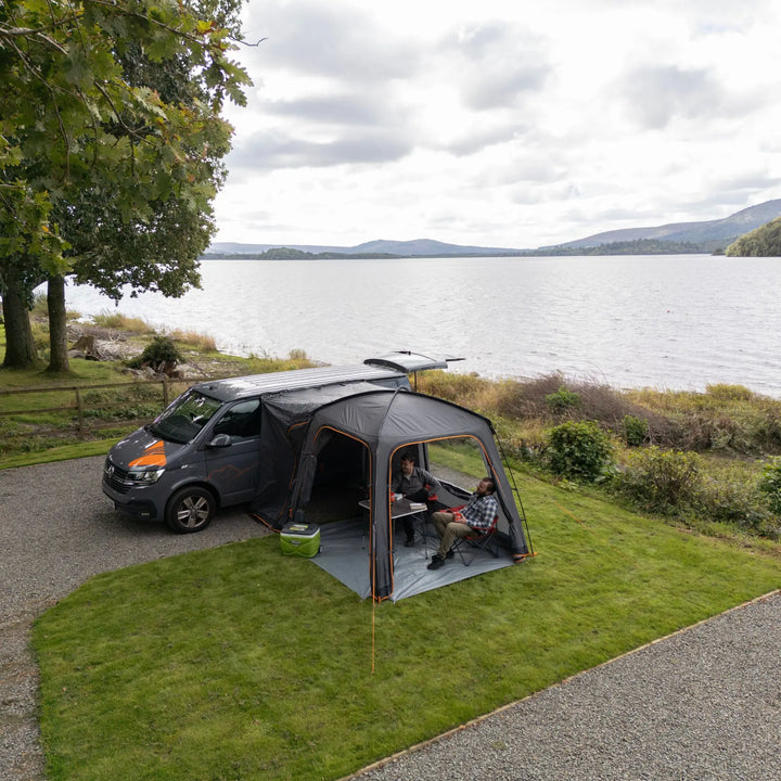 The Vango Tailgate Hub II Low awning connected to a campervan, with two campers seated inside enjoying the space and view.