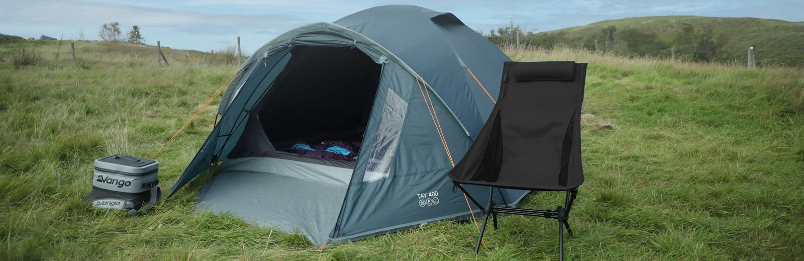 Wide view of the Vango Tay 400 4-man tent fully set up at a campsite with festival essentials like a Vango cooler and a camping chair in the foreground.