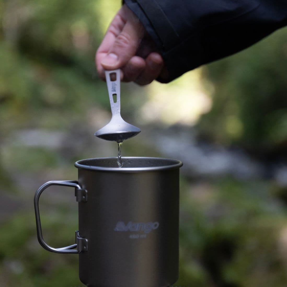 Hand holding the Vango Titanium spoon, dripping water into a Vango camping mug. Compact and lightweight camping cutlery for outdoor cooking.