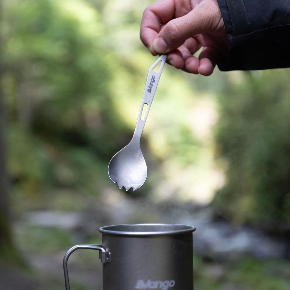 Person holding Vango Titanium Spork with opener above a Vango camping mug, ideal lightweight camping utensil.