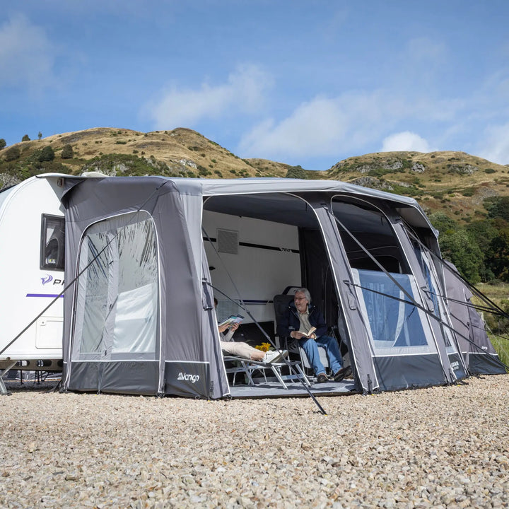 A Vango Tuscany Pro Air 400 Caravan Awning attached to a white caravan on a gravel campsite, with a man sitting inside under an open canopy. Rolling hills and blue sky are visible in the background.