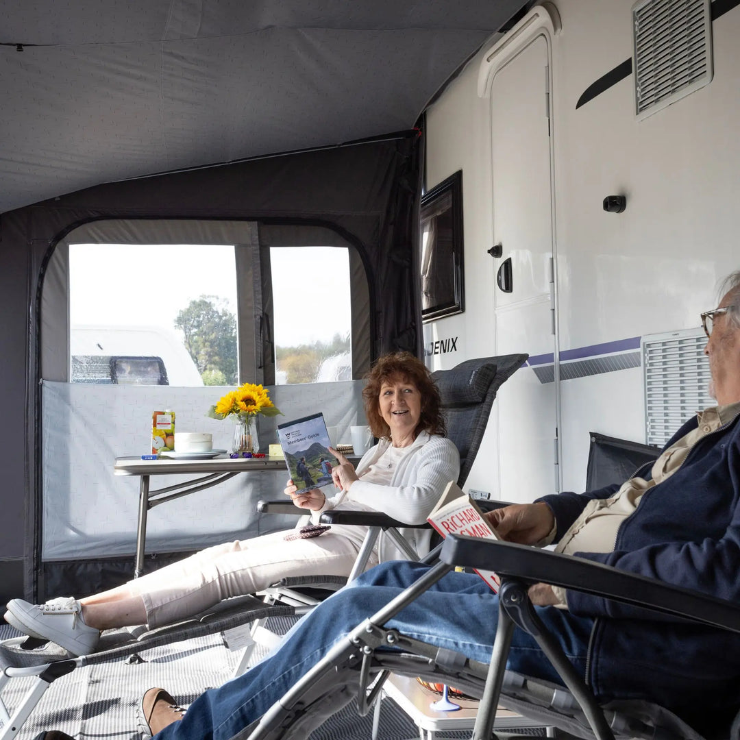 An older woman sits in a lounge chair inside a Vango Tuscany Pro Air 400 Caravan Awning, reading a book, while an older man sits in the foreground also reading. The scene is set in a bright, modern caravan.